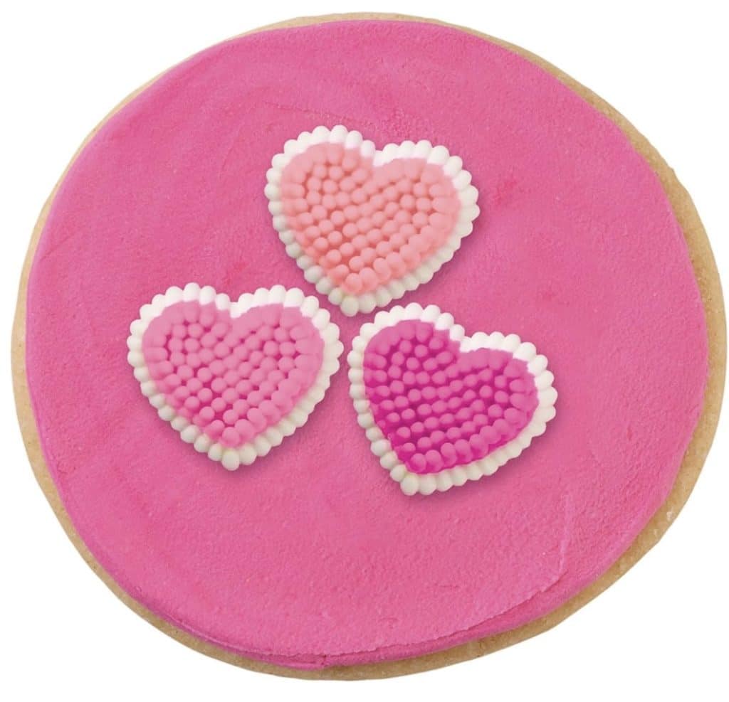 A round Valentine's Day cookie with pink icing is adorned with three heart-shaped toppings. The hearts, in various shades of pink, boast a beaded texture reminiscent of Rice Krispie Treats.